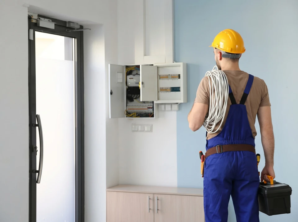 Électricien professionnel en salopette bleue et casque jaune dans cuisine moderne, portant câbles électriques et caisse à outils